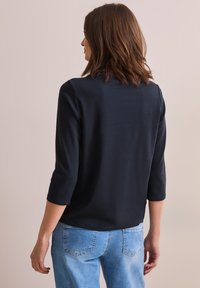 Top a maniche lunghe di colore blu navy con scollatura a barchetta, realizzato in tessuto morbido. Dotato di maniche a tre quarti e orlo curvo, indossato con jeans di denim chiaro.