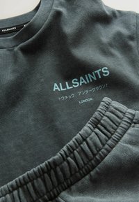 Hiilenharmaa huppari, jossa on teksti "ALLSAINTS" vaaleansinisellä. Sisältää ribatut pyöreät kaula-aukot ja sileän kankaan. Lähellä on yhteensopiva joustava vyötärö.