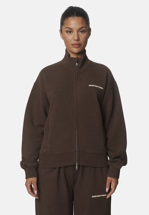 Donna con felpa marrone scuro con cerniera e pantaloni abbinati con loghi di testo bianchi, in piedi davanti a uno sfondo chiaro e uniforme.