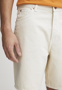 Pantalones cortos de algodón color beige claro con un ajuste relajado, que presentan herrajes en tono cobre y bolsillos frontales. La tela tiene un aspecto ligeramente texturizado.