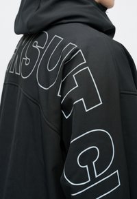 En person som har på seg en svart hettegenser med store, hvite bokstaver tegnet ut over ryggen og nedover ermet.