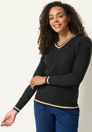 Vrouw met krullend haar die een zwarte V-hals trui met beige bies en een blauwe spijkerbroek draagt, glimlachend tegen een effen lichte achtergrond.