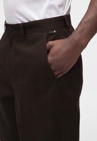 Pantalon marron foncé avec une main dans la poche avant, porté avec une chemise blanche visible à la taille, vu de côté.