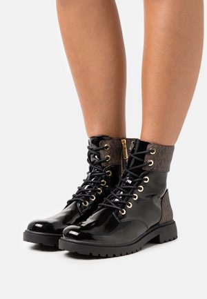 Bottes à lacets en cuir verni noir avec poignets en tissu marron et œillets dorés portées sur des jambes nues.
