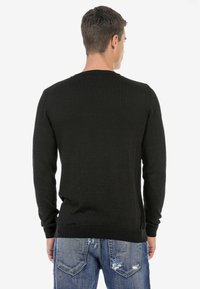 Schwarzer, langärmeliger Pullover mit geripptem Muster, Rundhalsausschnitt und eng anliegenden Bündchen, getragen über abgewetzten blauen Jeans.