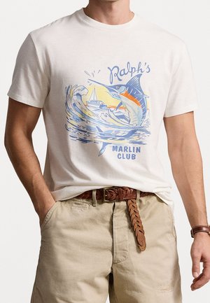 Mann trägt weißes T-Shirt mit blau-orangem Marlinfisch- und Boots-Design und beige Hose mit geflochtenem braunem Ledergürtel.
