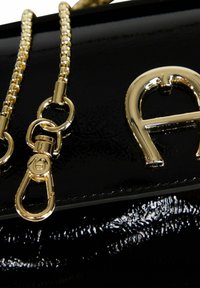 Borsa nera in vernice con tracolla a catena dorata, dettagli metallici e un logo dorato prominente sul davanti.