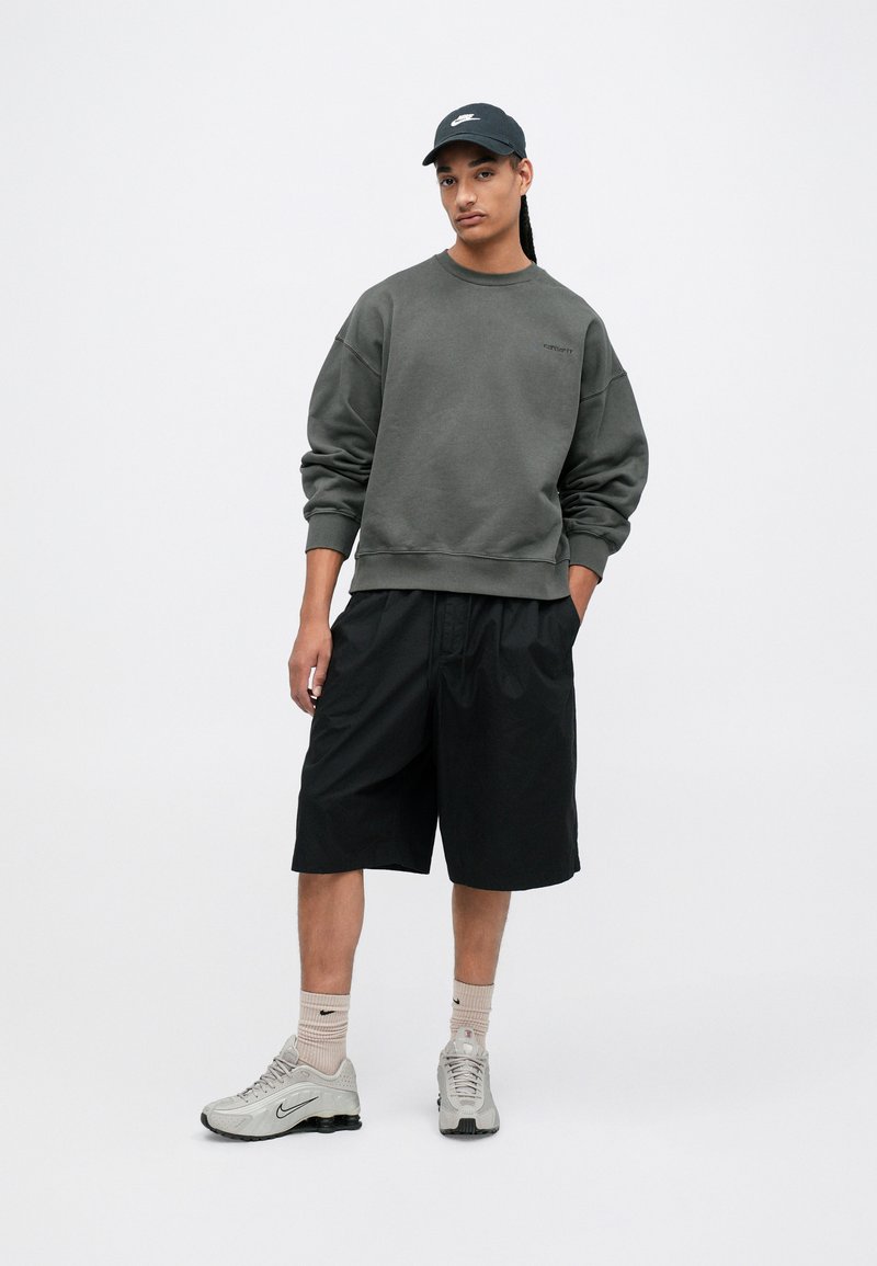 Sudadera gris, pantalones cortos negros, zapatillas grises, gorra negra. La sudadera tiene un cuello redondo; los pantalones cortos son holgados y de longitud a la rodilla.