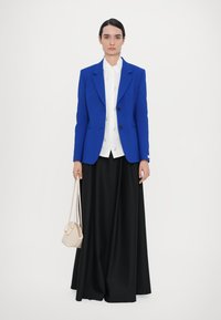 Blue tailored blazer, white shirt with printed details, flowing black skirt, and a small woven beige handbag. Structured and elegant outfit.