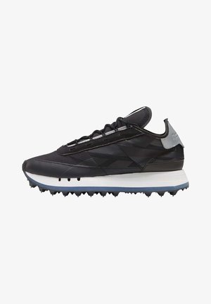 Zapatilla deportiva negra de corte bajo con parte superior texturizada, media suela blanca, suela azul con pinchos y lengüeta gris en el talón, mostrada de perfil.