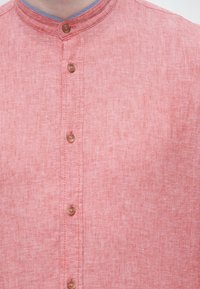 Κοντινό πλάνο ενός ατόμου που φοράει ροζ λινό πουκάμισο με μανδραρινένιο γιακά και τέσσερα ορατά ξύλινα κουμπιά στο εμπρός μέρος.