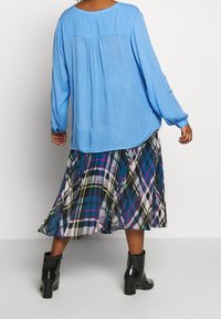 Vrouw in een blauwe blouse met lange mouwen, een meerkleurige geruite geplooide rok en zwarte haklaarzen die tegen een effen witte achtergrond staat.