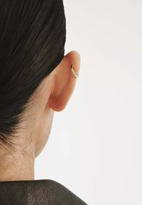 Pendiente geométrico plateado en forma de V, colocado en una oreja perforada. Superficie lisa con un diseño minimalista, complementando el cabello oscuro y el fondo.