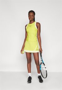 Top deportivo amarillo sin mangas y falda con el logo de Nike en negro, acompañados de una raqueta y una pelota de tenis. La falda presenta un diseño en capas con acentos blancos.