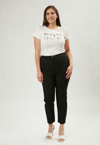 Witte t-shirt met kantmouwen en grafische tekst, gecombineerd met zwarte strakke broeken en witte hakken, staand op een neutrale achtergrond.