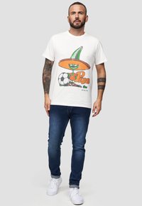 Hvid bomulds T-shirt med en tegneseriefigur iført en grøn sombrero og orange detaljer, ved siden af en fodbold. Båret med blå jeans og hvide sneakers.