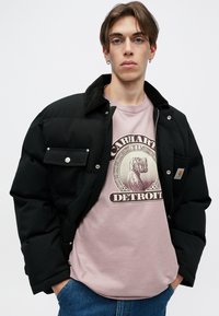 Chaqueta puffer negra con botones a presión y dos bolsillos frontales, llevada sobre una camiseta gráfica rosa clara con un diseño de moneda, combinada con jeans azules.