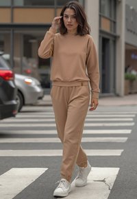 Beige sportos szett hosszú ujjú, kerek nyakú felsővel és szűkített jogger nadrággal. Rugalmas mandzsettákkal és derékrésszel rendelkezik, fehér sportcipőkkel párosítva.