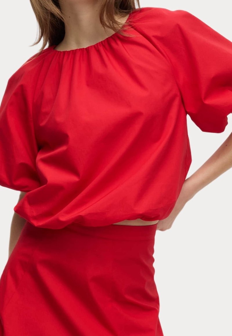 Femme portant un chemisier rouge vif à manches bouffantes avec un col froncé et une jupe taille haute assortie, debout avec une main sur la hanche.