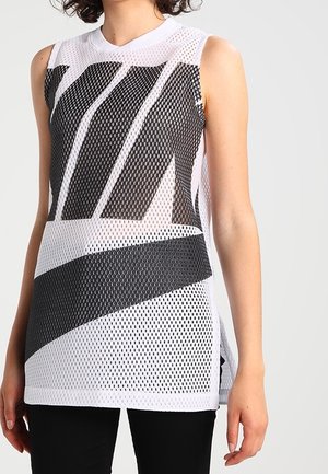 Haut sans manches en maille blanche avec de grandes rayures abstraites noires porté sur des vêtements noirs, sur une personne devant un fond uni.