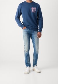 Granatowa bluza z różowym nadrukiem graficznym, zestawiona z jasnoniebieskimi wyblakłymi dżinsami i białymi trampkami. Casualowy, luźny fason z ściągaczami na końcach rękawów.