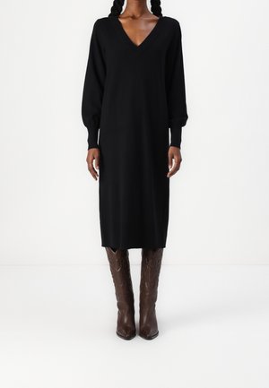 Femme portant une longue robe noire à encolure en V avec des manches ballon, associée à des bottes de cowboy en cuir marron montant jusqu'au genou.