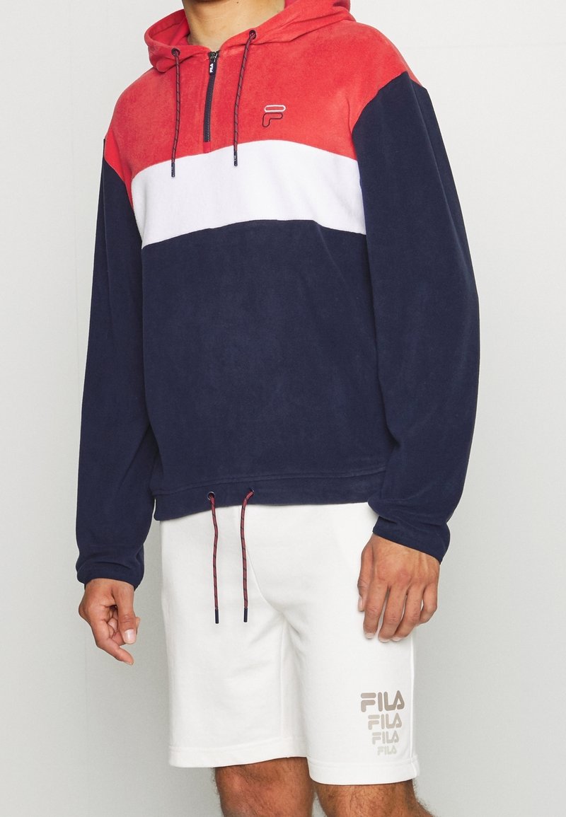 Pull-over à capuche en polaire rouge, blanc et bleu marine avec un zip frontal demi-long. Il présente un cordon de serrage réglable et un logo sur la poitrine. Associé à un short blanc.