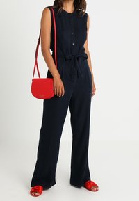 Námornický overal bez rukávů, s předními knoflíky a vázaným pasem, doplněný červenou příčnou taškou a červenými sandály na otevřenou špičku.