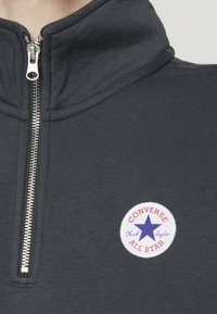 Sudadera con cremallera de color gris oscuro y cuello alto, con un parche redondo blanco que dice "Converse All Star" y un logo de estrella azul.
