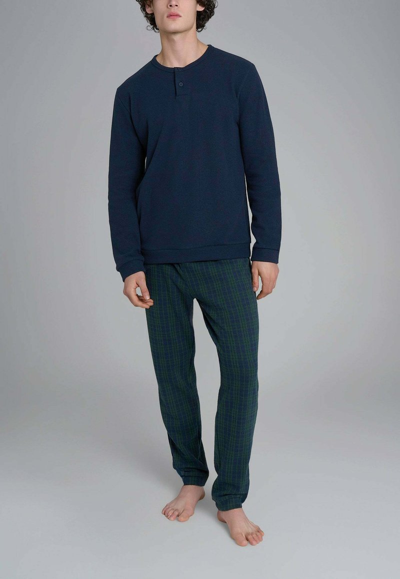 Giovane uomo in piedi a piedi nudi che indossa una maglia henley a maniche lunghe blu navy e pantaloni del pigiama a quadri verde scuro su sfondo semplice.
