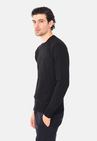 Pullover nero a maniche lunghe con design aderente, dotato di polsini e orlo a coste, realizzato in un materiale liscio e morbido. Indossato sopra pantaloni scuri.