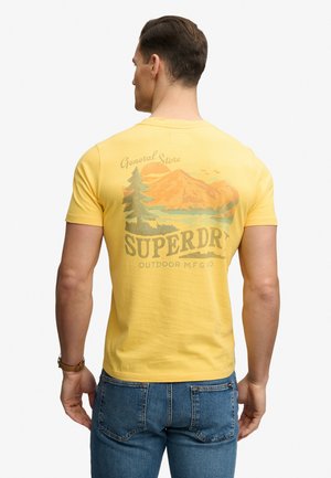 Uomo che indossa una maglietta gialla con grafica di montagna, albero e lago e la scritta "Superdry Outdoor M.F.G Co", abbinata a jeans blu.