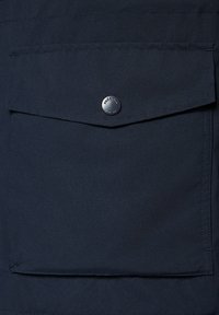 Granatowy materiał o gładkiej fakturze, z kieszenią na klapkę zabezpieczoną okrągłym srebrnym guzikiem. Minimalistyczny design z czystymi liniami.