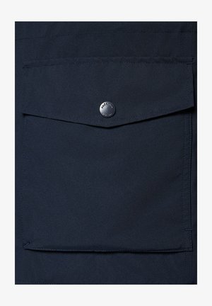 Granatowy materiał o gładkiej fakturze, z kieszenią na klapkę zabezpieczoną okrągłym srebrnym guzikiem. Minimalistyczny design z czystymi liniami.