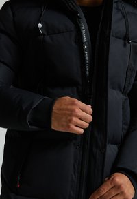 Svart polstret jakke med glidelås foran, med elastiske mansjetter og en fremtredende merkelapp inni kragen. Myk, glatt tekstur.