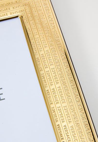 Foto rāmis zelta rāmītī ar reljefa "Versace" logotipu, gludu, spīdīgu virsmu un taisnstūrveida formu. Iekšpusē atrodas balts ieliktnis.