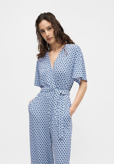 Γυναίκα με κυματιστά μαλλιά που φοράει ένα μπλε και άσπρο ολόσωμο κοντομάνικο με γεωμετρικό μοτίβο, με δεμένη ζώνη στη μέση και χέρια στις τσέπες.