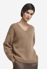 Pull beige en tricot avec encolure en V, coupe ample et poignets et ourlet côtelés. Texture douce et design décontracté idéal pour être superposé.