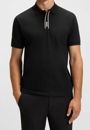 Homme portant un polo noir à manches courtes avec une étiquette "BOSS" sur la patte de boutonnage, associé à un pantalon noir, mains détendues le long du corps.