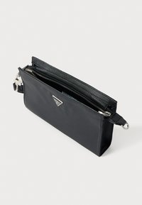 Μαύρο τσαντάκι χειρός από νάιλον με διακοσμητικό λογότυπο σε ασημί τόνο, κλείσιμο με φερμουάρ και αποσπώμενο λουράκι. Λείο υλικό, ορθογώνιο σχήμα, μινιμαλιστικός σχεδιασμός.