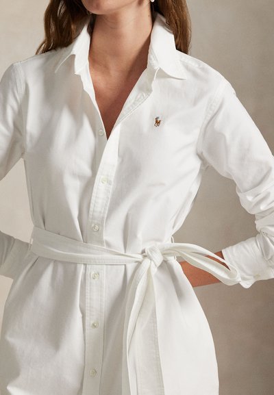 Nő, aki fehér, hosszú ujjú, patentos ingruhát visel gallérral és derékban kötött övvel, melyen a mellrészen kis hímzett logó található.