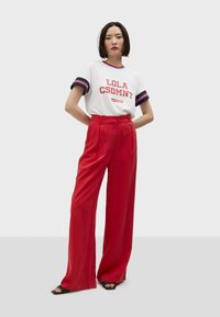 White t-shirt with red and blue accents featuring "LOLA CSDMNT" text, paired with high-waisted red wide-leg pants and black sandals.