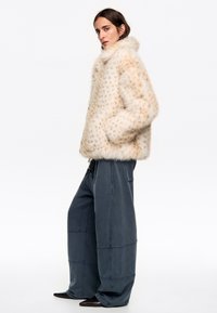 Chaqueta de pelaje sintético en beige con patrón de manchas, que presenta un cuello alto y bolsillos laterales. Combinada con pantalones grandes de color azul oscuro.