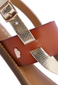 Sandal de cuero marrón con correas metálicas de plata texturizada y un hebilla decorativa dorada. Presenta un pequeño acento con forma en el lateral.