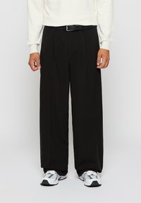 Mand iført sorte bukser med brede ben, hvid langærmet skjorte, sort bælte og hvide sneakers, stående mod en ensfarvet baggrund.