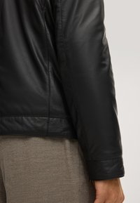 Schwarze Lederjacke mit glatter Textur, die eine strukturierte Passform aufweist, sichtbare Nähte hat und einen sauberen Abschluss am Saum und an den Ärmeln bietet.