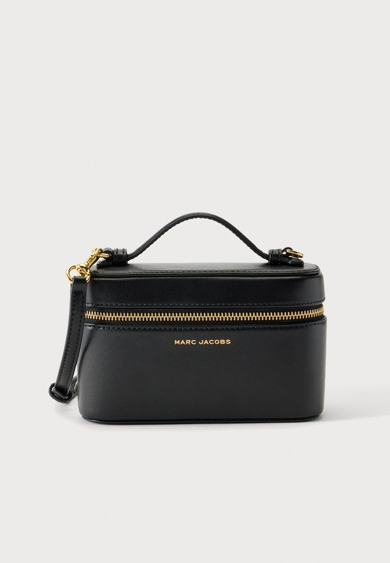 Μικρή δομημένη μαύρη τσάντα χειρός Marc Jacobs με χρυσό φερμουάρ, κοντή λαβή, αποσπώμενο λουράκι και χρυσό λογότυπο μπροστά, σε λευκό φόντο.