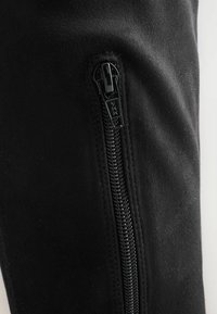 Schwarzer Wildlederstoff mit einem sichtbaren Reißverschluss. Der Reißverschluss ist aus schwarzem Metall, trägt ein YKK-Logo und verläuft an der Seite des Artikels.