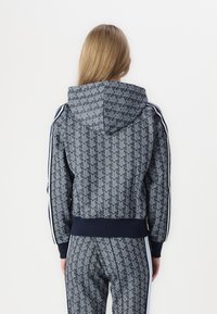 Námožná športová bunda s kapucňou v námorníckej modrej farbe s geometrickým vzorom. Obsahuje skrátený strih, rebrovaný lem a biele bočné pruhy na rukávoch.