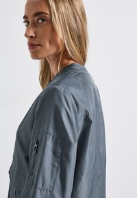 Veste bomber grise au tissu lisse, avec un col côtelé et une poche latérale munie d'une fermeture éclair. Design simple, coupe ajustée, sans motifs visibles.